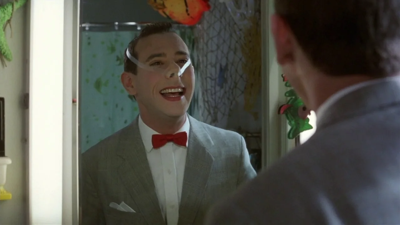 Pee-Wee’s Big Adventure (1985) movie still: medium — A man in a grey suit and red bow tie smiles at his reflection. He wears white adhesive tape…