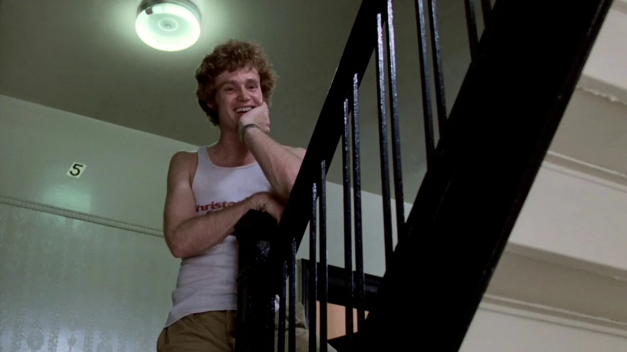 Cruising (1980) movie still: medium — A residential apartment building stairwell features a wall marked with the number five. A young man in a white…