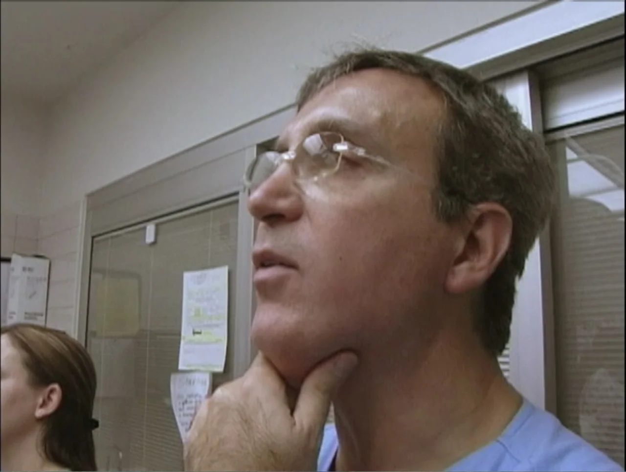 Addiction (2024) movie still: closeup — A clinical room with beige walls and documents taped to a glass-paneled door. A man in blue scrubs stands in…