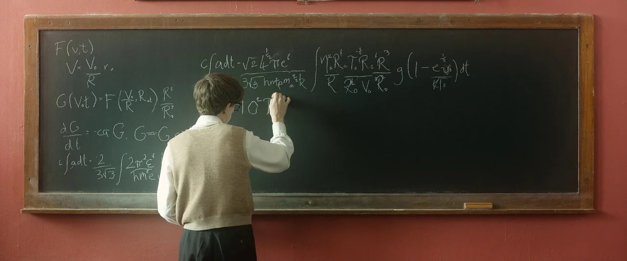 The Theory of Everything (2014) movie still: medium — A classroom with a dark blackboard mounted on a deep red wall. A young man wearing a tan vest…