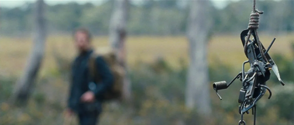 The Hunter (2011) movie still: closeup — A metal animal trap hangs suspended from a rope in a wooded clearing. A blurred man with a backpack stands…
