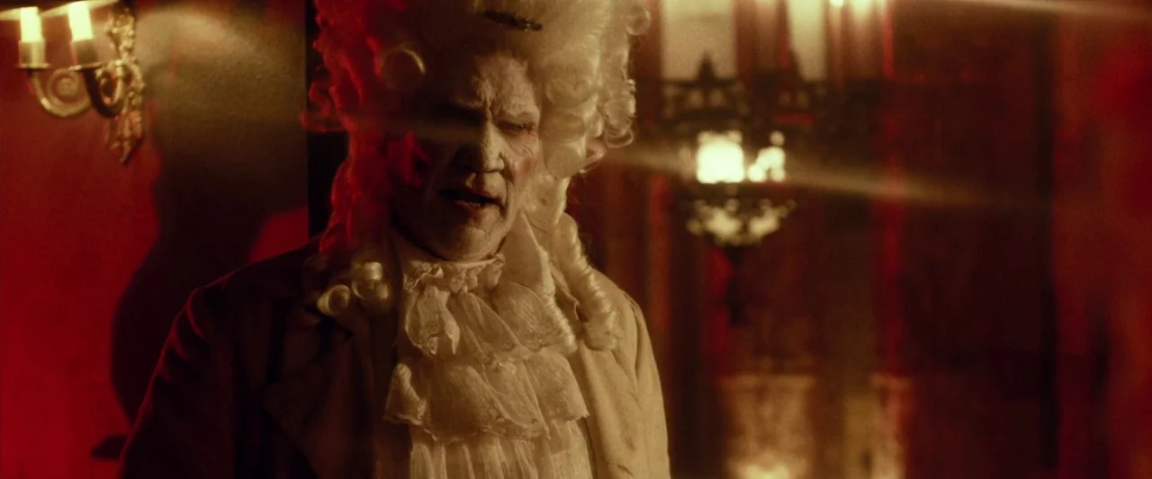 31 (2016) movie still: medium — A dimly lit room with red walls and decorative wall sconces. A man in an 18th-century costume stands in the…