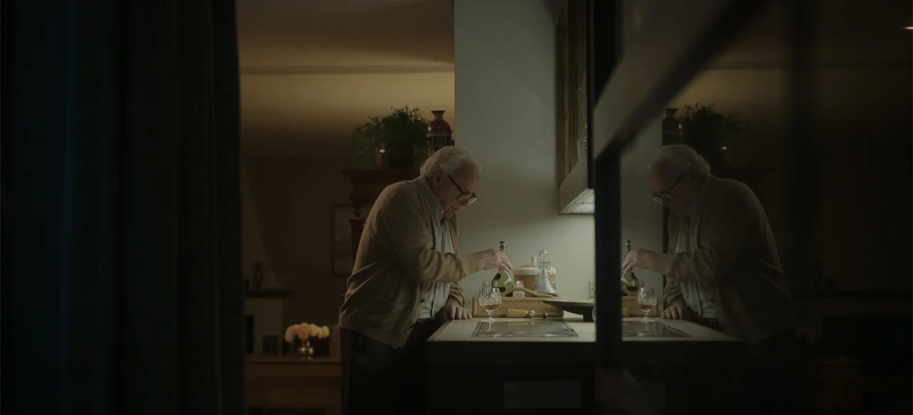 One Life (2023) movie still: medium — A dimly lit kitchen features a counter with a bottle and a glass. An elderly man pours a drink while standing…