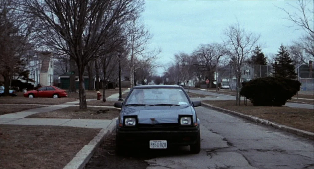 Buffalo ’66 (1998) movie still: wide — A dark sedan is parked on the edge of a paved suburban street. Leafless trees and houses line the sidewalk…