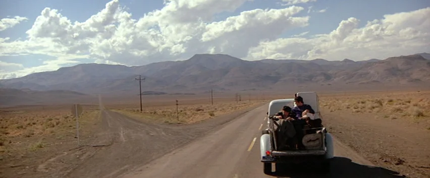 Scarecrow (1973) movie still: wide — An arid, sprawling desert plain surrounds an empty paved road extending to the mountains. Two men sit inside…