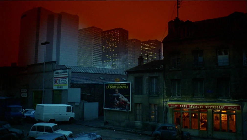 The American Friend (1977) movie still: establishing — A dark city street at night features a restaurant and low-rise buildings against skyscrapers.…