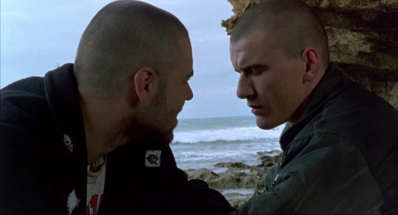 Romper Stomper (1992) movie still: medium — A rocky beach shoreline under an overcast sky by the ocean waves. Two men with buzzed hair face each…