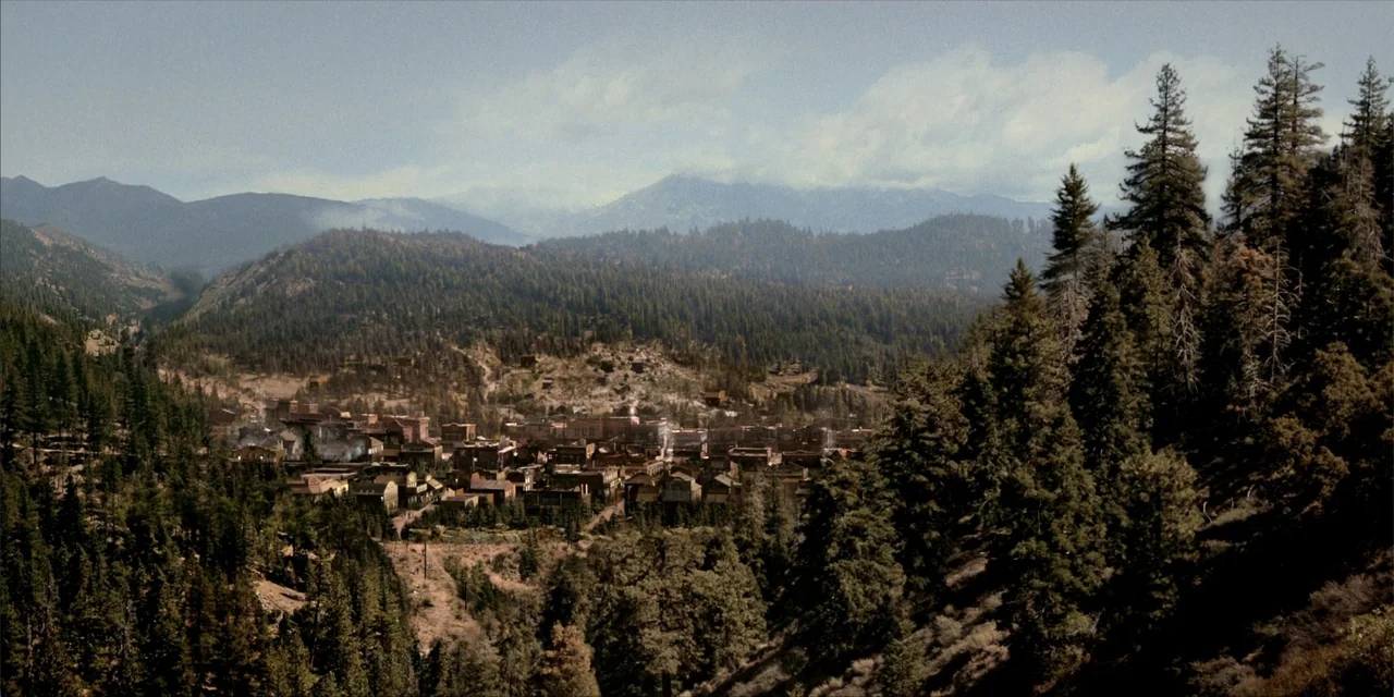 Deadwood: The Movie (2019) movie still: establishing — A remote mountain town sits nestled within a dense forest of evergreen trees. Mountains rise…