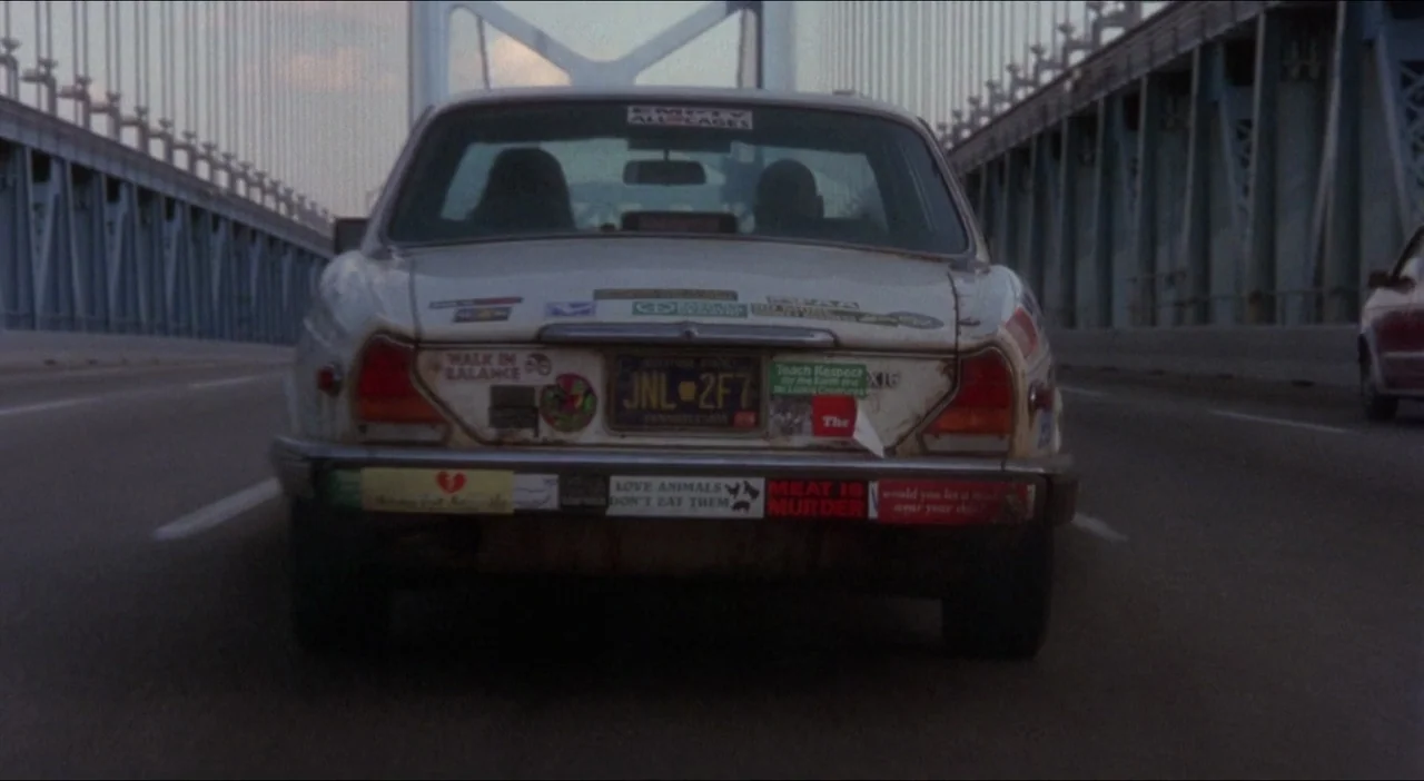 Twelve Monkeys (1995) movie still: medium — A white sedan covered in bumper stickers travels across a large metal suspension bridge. The car shows…