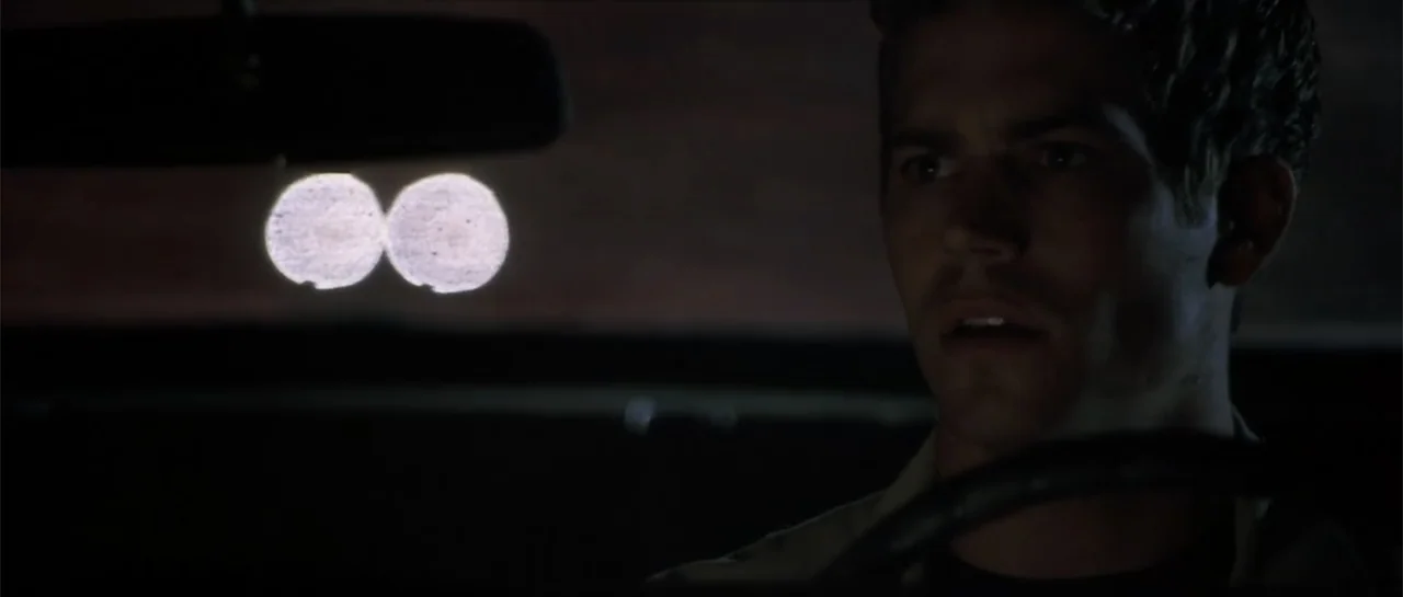 Joy Ride (2001) movie still: medium — A man sits in the driver seat of a vehicle at night. Two bright circles of light from oncoming headlights…