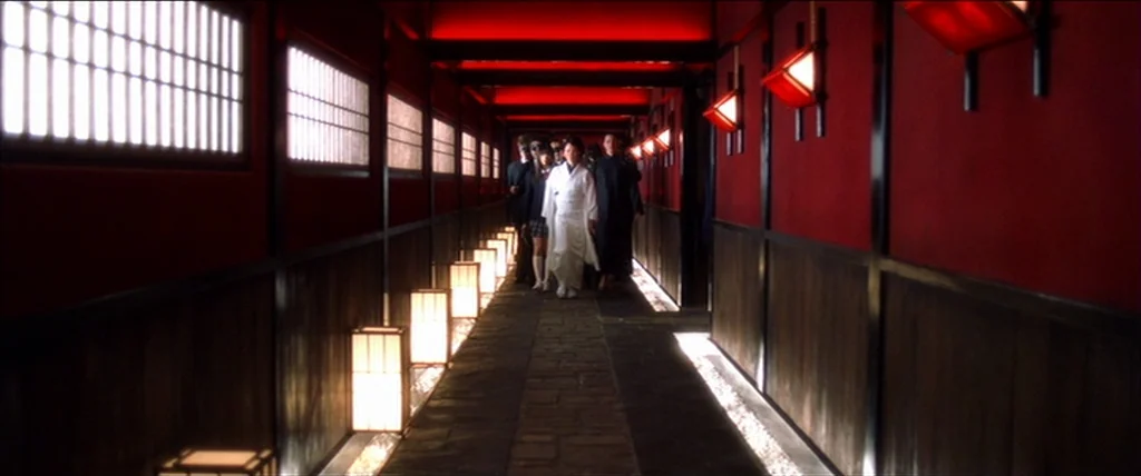 Kill Bill Vol. 1 (2004) movie still: wide — A long indoor hallway with deep red walls and dark wood panels features floor lanterns. A group led by a…