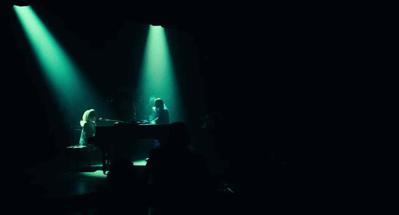 Joker: Folie à Deux (2024) movie still: wide — A dark, cavernous music venue is lit only by intense, narrow green overhead beams. Two performers sit…