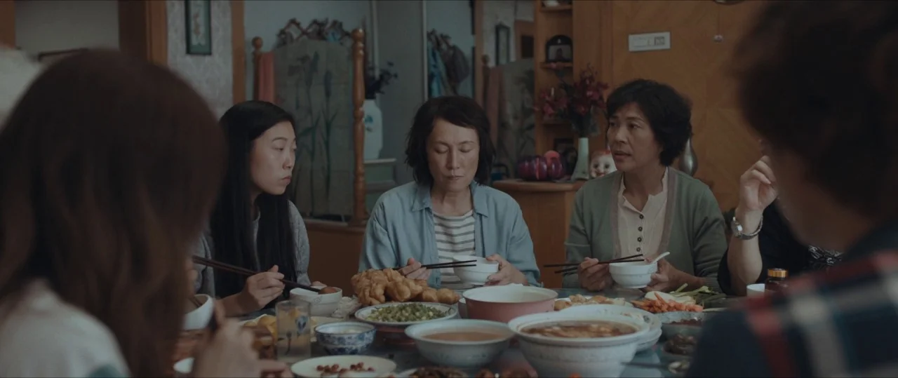 The Farewell (2019) movie still: medium — A family sits around a table full of bowls with Chinese cuisine. They hold chopsticks while eating in a…