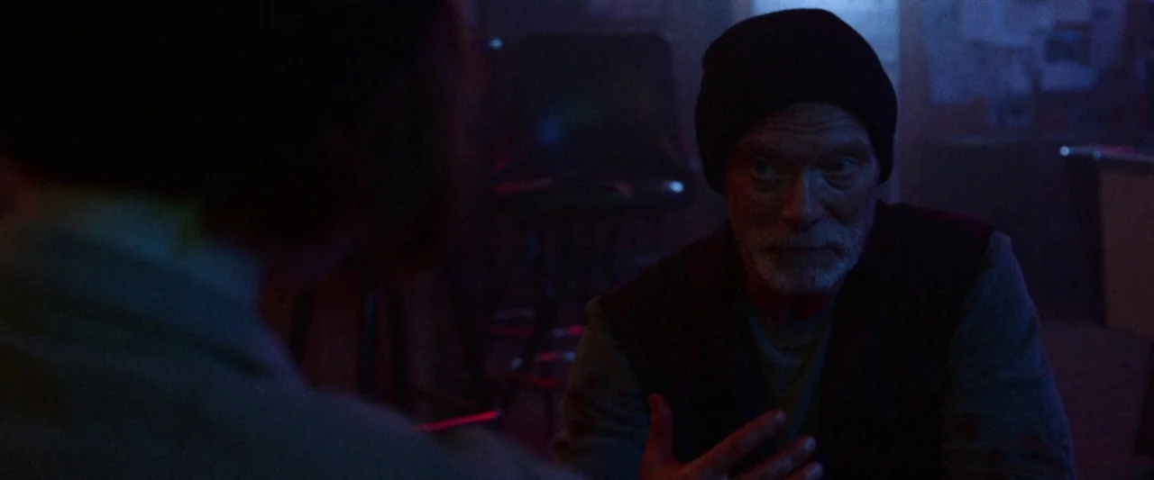 VFW movie still: medium — A dimly lit interior space with dark walls and scattered furniture. An older man in a beanie gestures with his hand during…