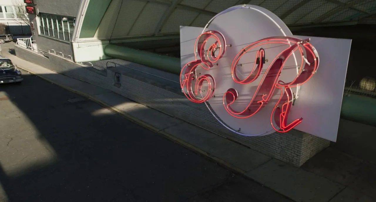 Motherless Brooklyn (2019) movie still: wide — A street scene under a concrete bridge featuring a prominent red neon E&R sign. A vintage car is…
