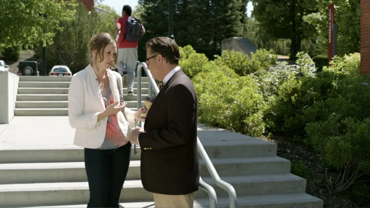 At Middleton (2013) movie still: medium — Concrete stairs outside a brick academic building with green foliage and paved paths. A woman gestures…