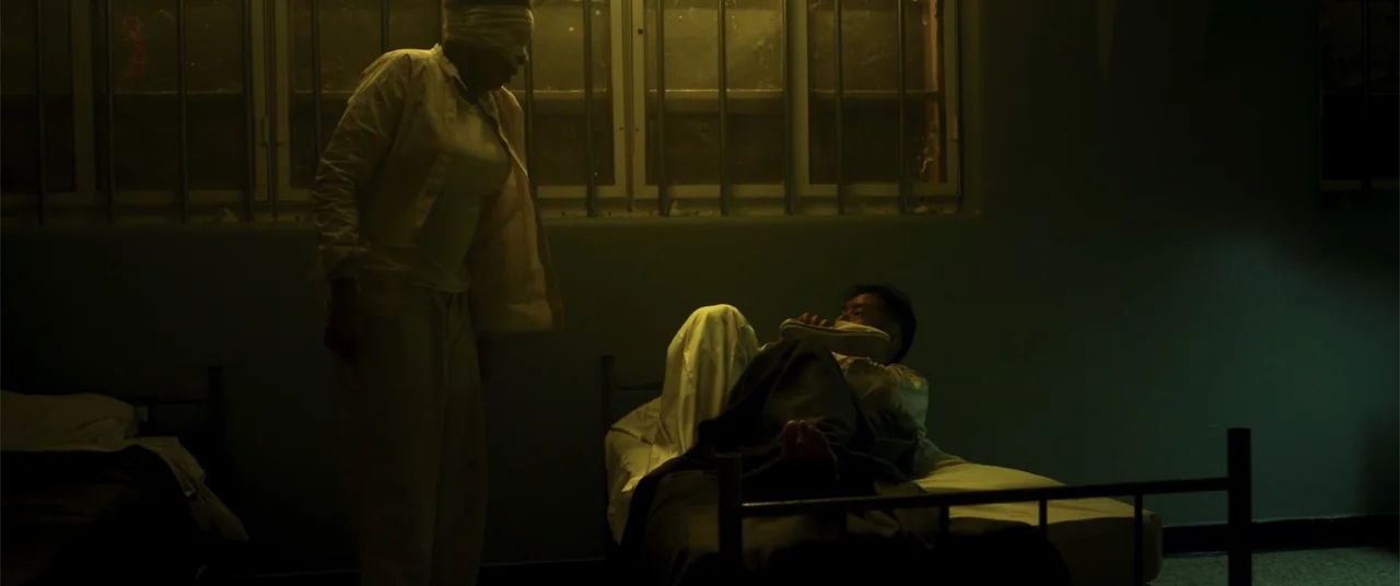 Phobias (2021) movie still: medium — A dimly lit room features a barred window and two simple cots with white bedding. One person stands in a…
