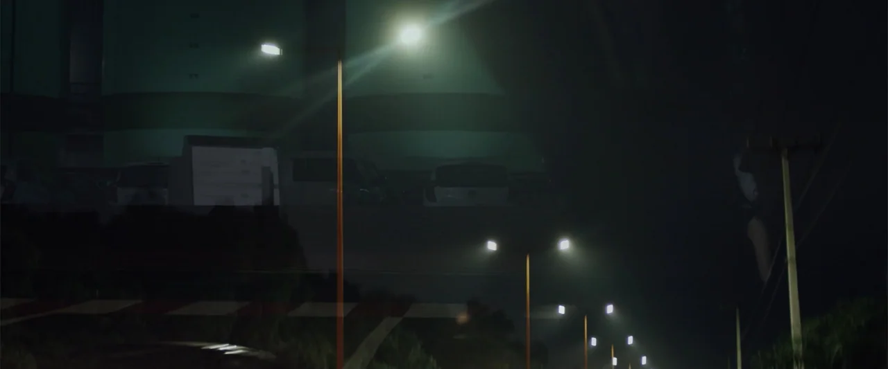 La Musiquita por Dentro (2019) movie still: wide — A dark road at night is illuminated by a row of streetlamps. Parked white cars sit beneath a…