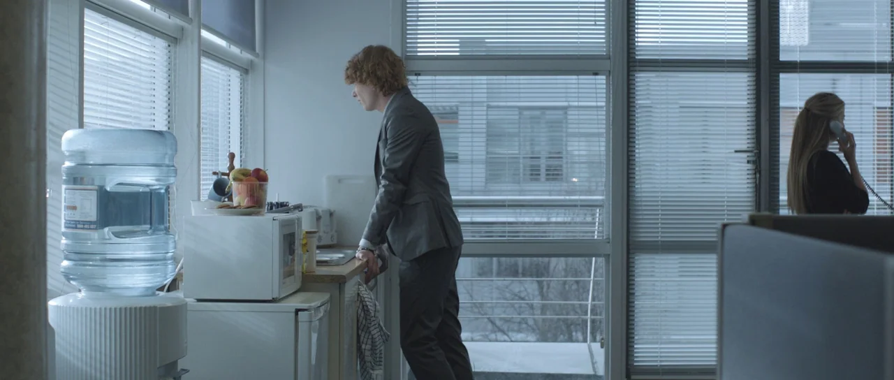 Frank (2014) movie still: wide — A modern workplace kitchenette features a water cooler, microwave, and windows with horizontal blinds. A red-haired…