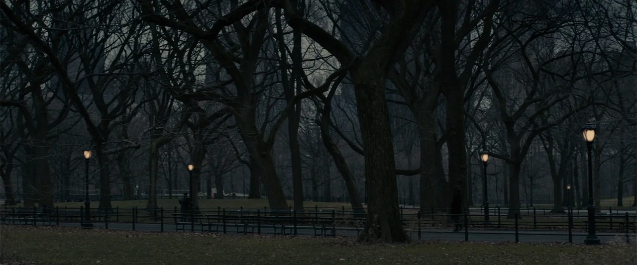 Winter's Tale (2014) movie still: wide — A dense urban park during dusk with numerous bare trees and dark pathways. Rows of wrought-iron benches are…
