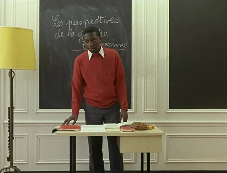 La Chinoise (1967) movie still: medium — A classroom with white wood-paneled walls and a black chalkboard with handwritten cursive text. A Black man…