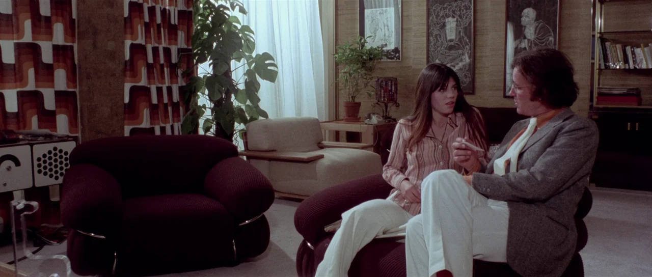 The Suspicious Death of a Minor (1975) movie still: medium — A 1970s living room features heavy geometric patterned curtains and dark fabric…