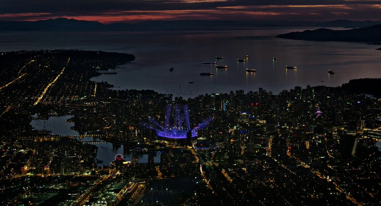 Annette (2021) movie still: establishing — An aerial nighttime view displays a densely lit coastal city bordering a dark ocean. A stadium centrally…