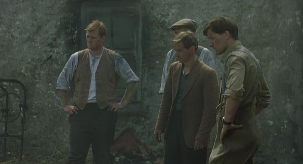 The Wind That Shakes the Barley (2006) movie still: fullbody — An exterior scene depicts four men standing in front of a rustic stone wall. They are…