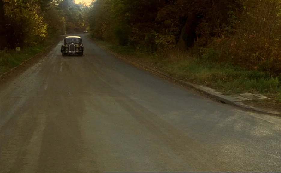 Murmur of the Heart (1971) movie still: wide — An asphalt road stretches through a wooded area lined with dense autumn trees. A vintage car drives…