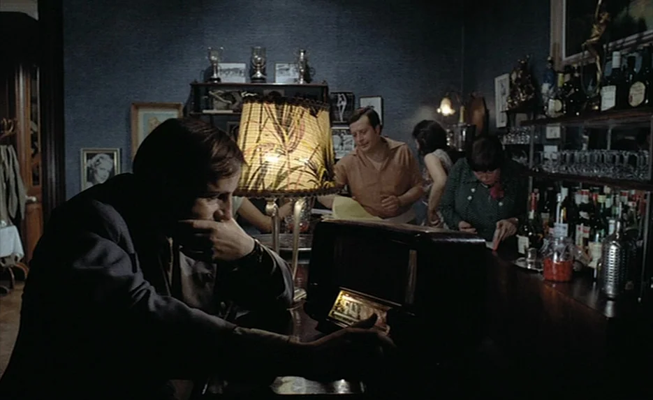 Lacombe, Lucien (1974) movie still: medium — A dimly lit room with dark wallpaper is filled with various framed pictures and trophies. A man sits in…