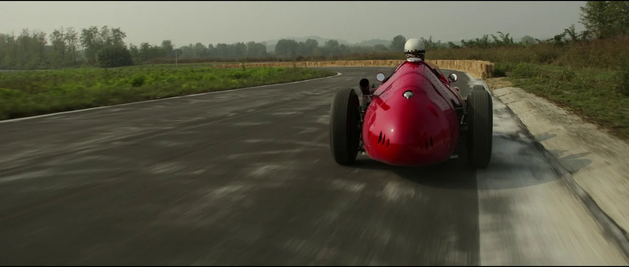 Ferrari (2023) movie still: wide — An asphalt racetrack curves through a hazy landscape featuring dry grass and distant trees. A red vintage…