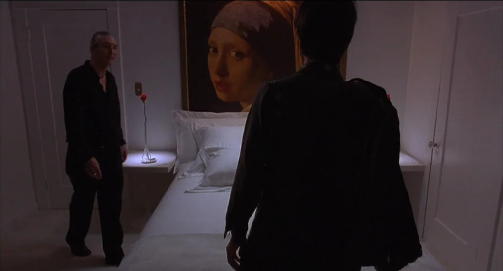 Mysterious Skin (2004) movie still: medium — A dimly lit room with white walls contains a bed and a large painting. An older man stands near a bed…