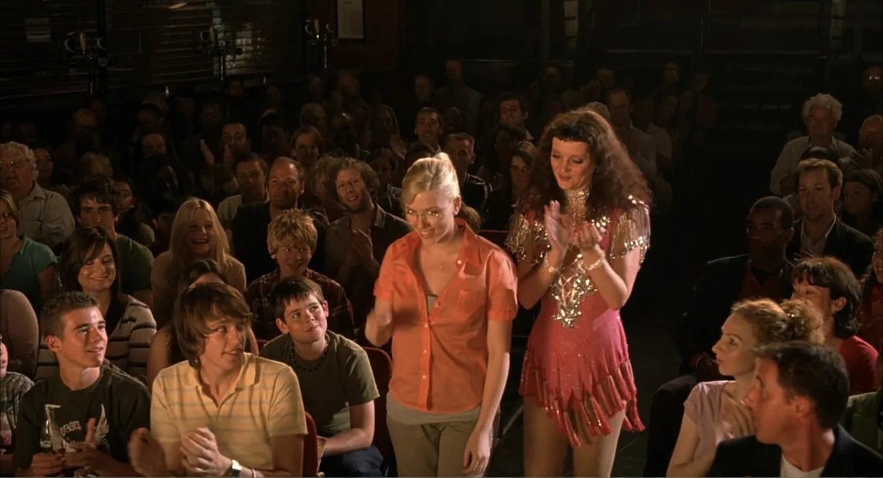 Scoop (2024) movie still: medium — An audience sits in a dimly lit hall watching a performance. Two women stand in the center aisle clapping while…