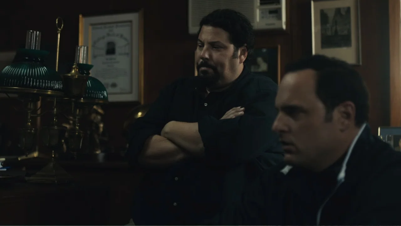 Paterno (2018) movie still: medium — A wood-paneled professional office features framed documents on the walls and a desk lamp. Two men in dark…
