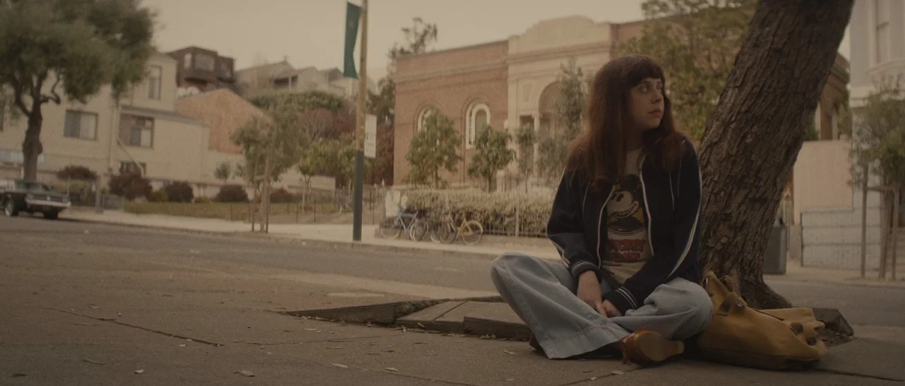 The Diary of a Teenage Girl (2015) movie still: medium — A quiet urban sidewalk features a woman resting against a tree trunk. Buildings and parked…