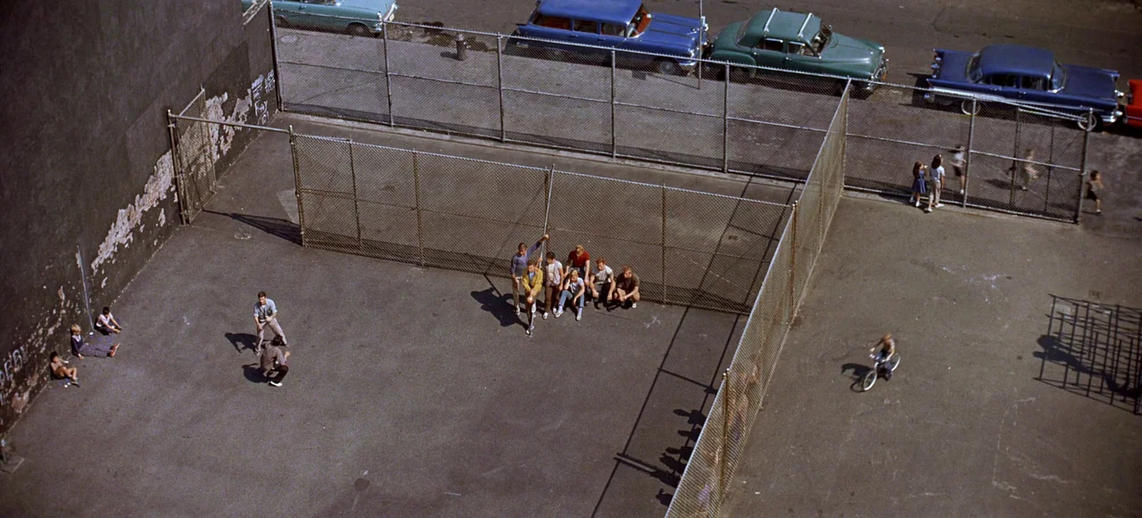 West Side Story (1961) movie still: establishing — An asphalt-surfaced outdoor area is enclosed by high chain-link fencing near parked vintage…