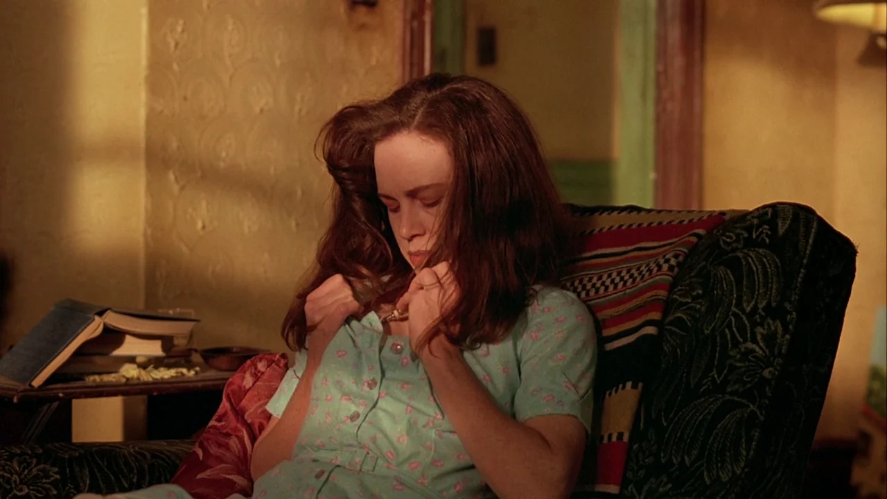 Naked Lunch (1991) movie still: medium — A dimly lit room with textured yellow walls and dark wooden furniture. A young woman sits in a patterned…