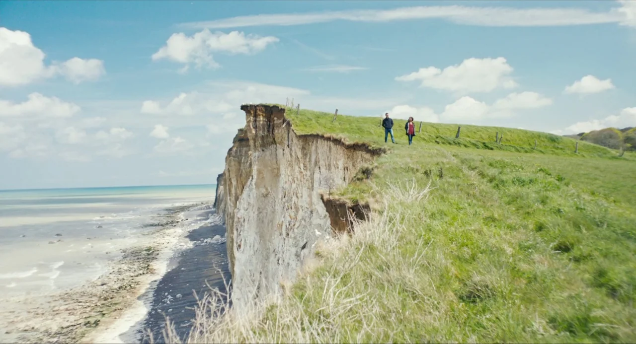 Submergence movie still: wide — A high, chalky cliff drops sharply into the ocean below a bright blue sky. A man and a woman walk along the grassy…