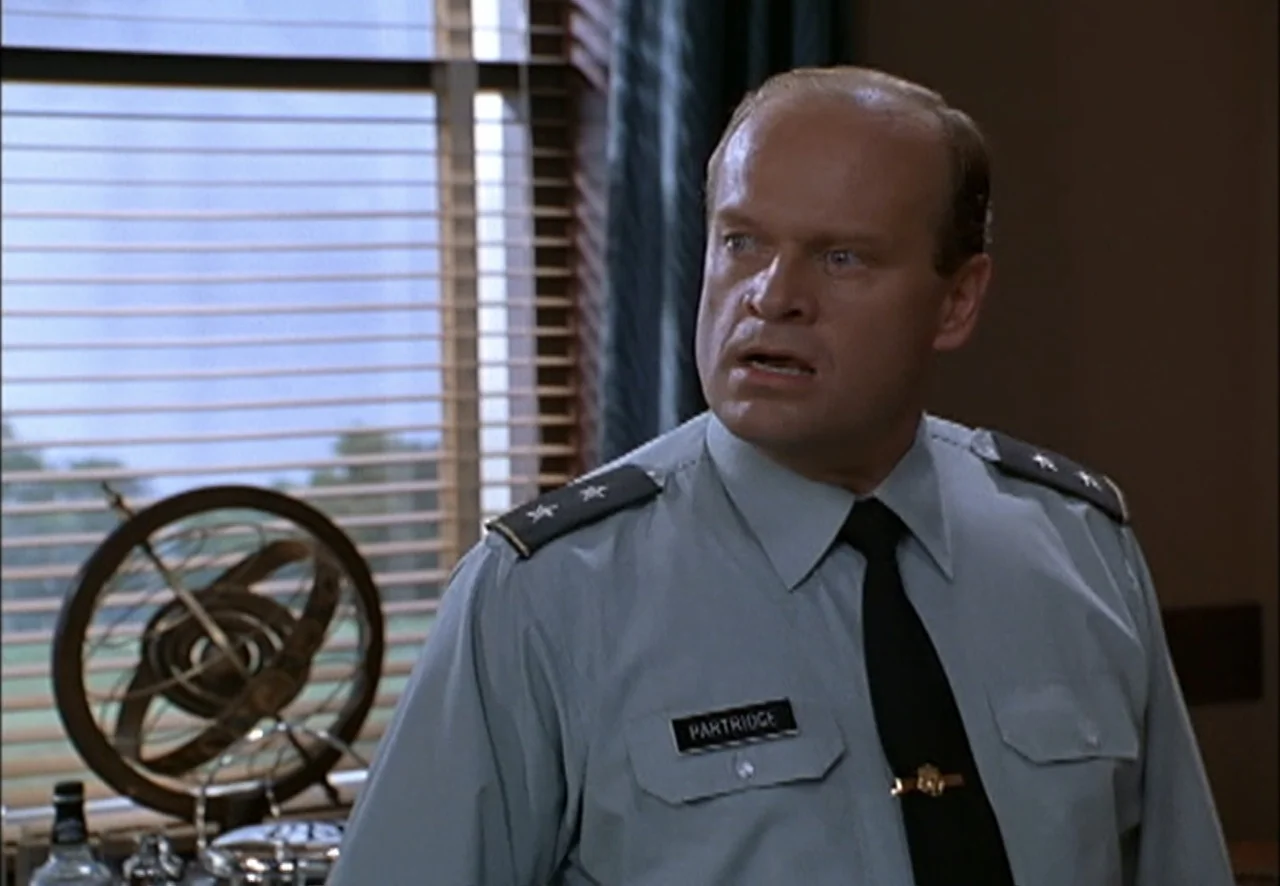The Pentagon Wars (1998) movie still: medium — An interior office with a window featuring closed blinds and a desk ornament. A man in a military…