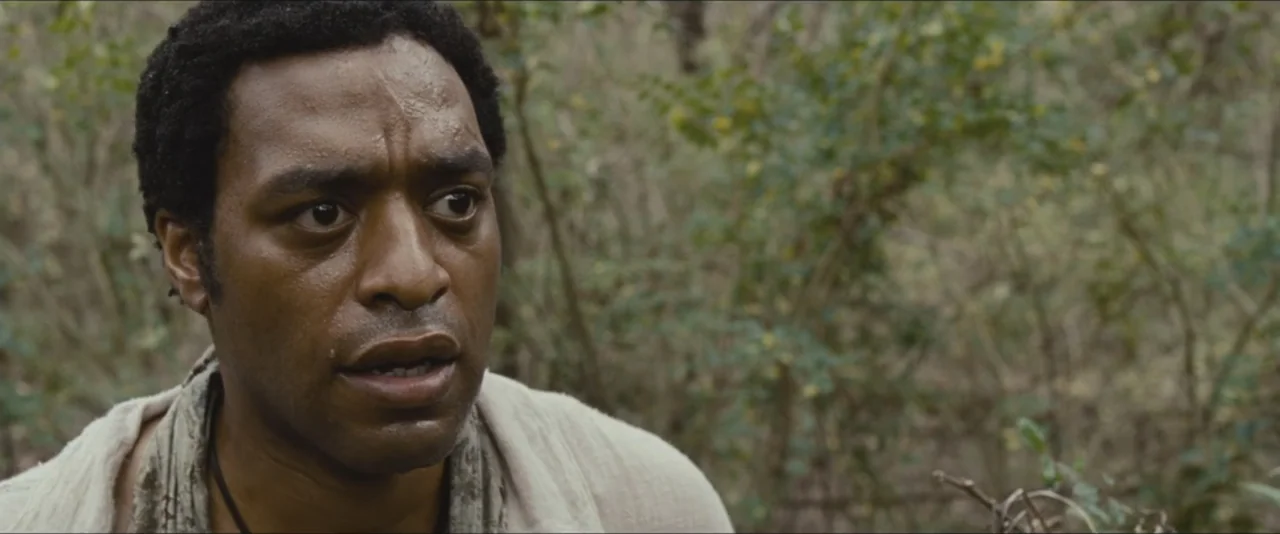 12 Years a Slave (2013) movie still: closeup — A dense woodland environment with blurred tree branches and foliage visible behind the subject. A man…