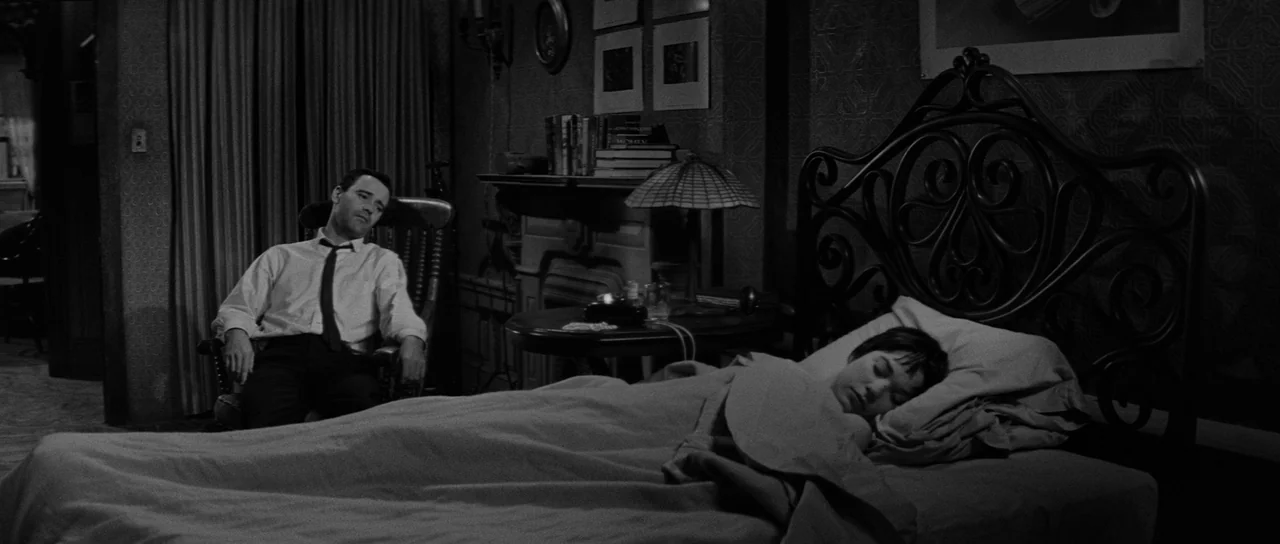 The Apartment (1960) movie still: wide — A 1960s furnished room features a decorative wrought iron bed frame and fireplace mantel. A man observes a…