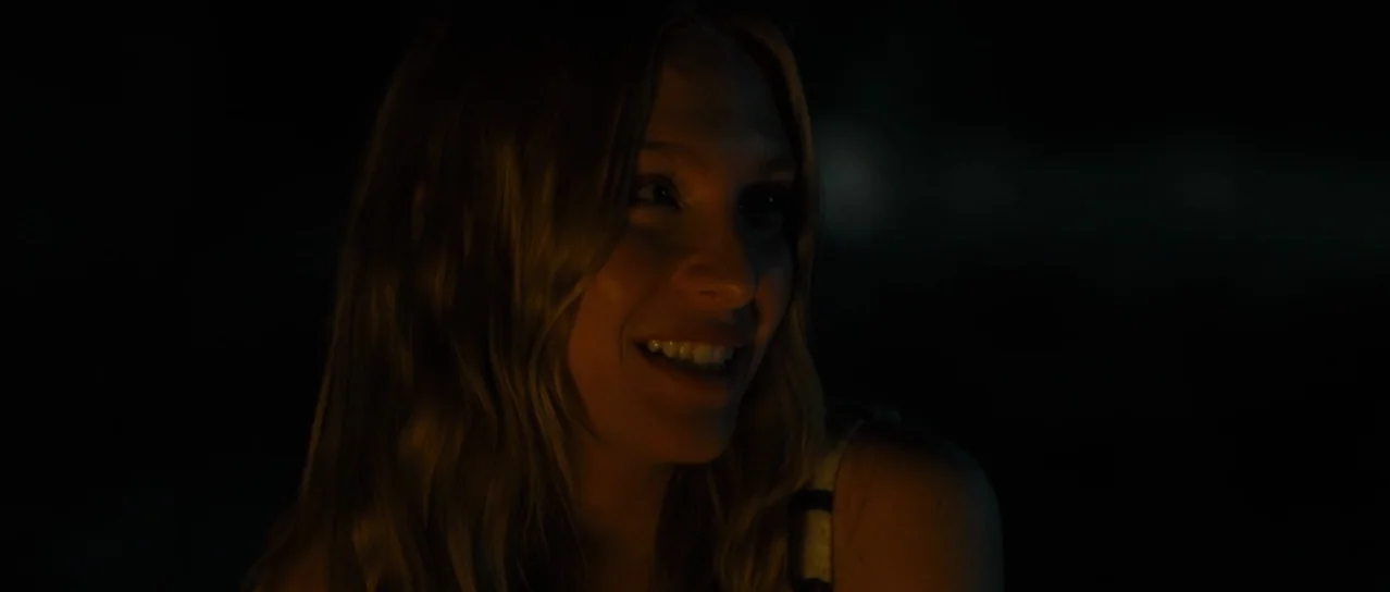 Road Games (2015) movie still: closeup — A dark outdoor space at night with only the subject's face clearly visible. The woman smiles while facing…