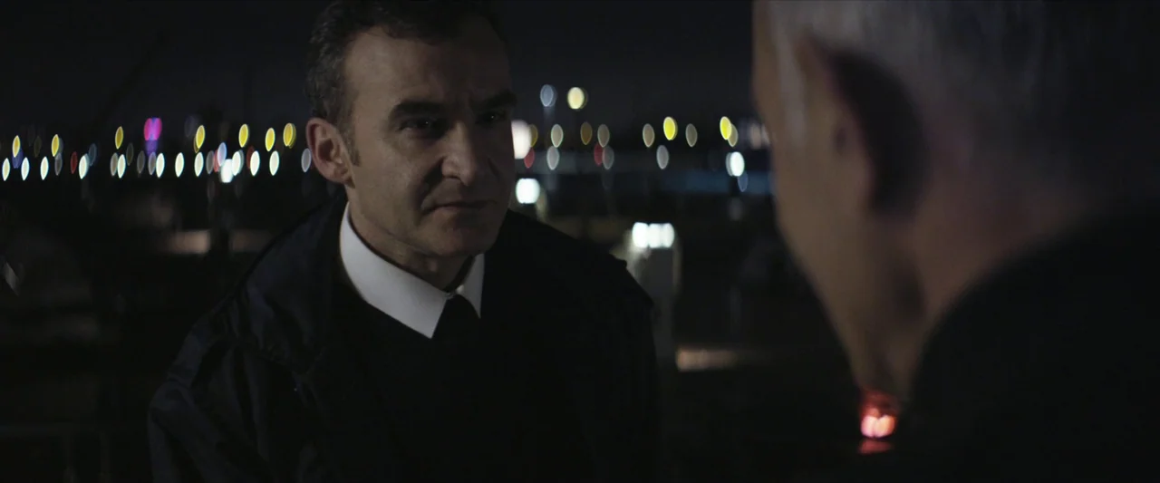 Chevalier (2015) movie still: medium — A night exterior setting near a water body with blurred city lights in background. A middle-aged man in a…