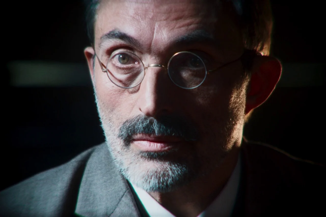 The Settlers (2023) movie still: closeup — A man stands in a dark environment with minimal visible background details. The subject faces forward…