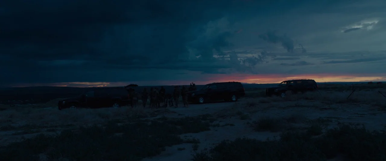 Sicario (2015) movie still: wide — A group of people stands between two dark SUVs on a flat desert terrain. The horizon shows a sunset with heavy…