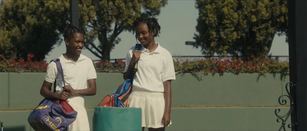 King Richard (2021) movie still: medium — An outdoor tennis court features a green perimeter wall and lush trees in background. Two teenage girls…