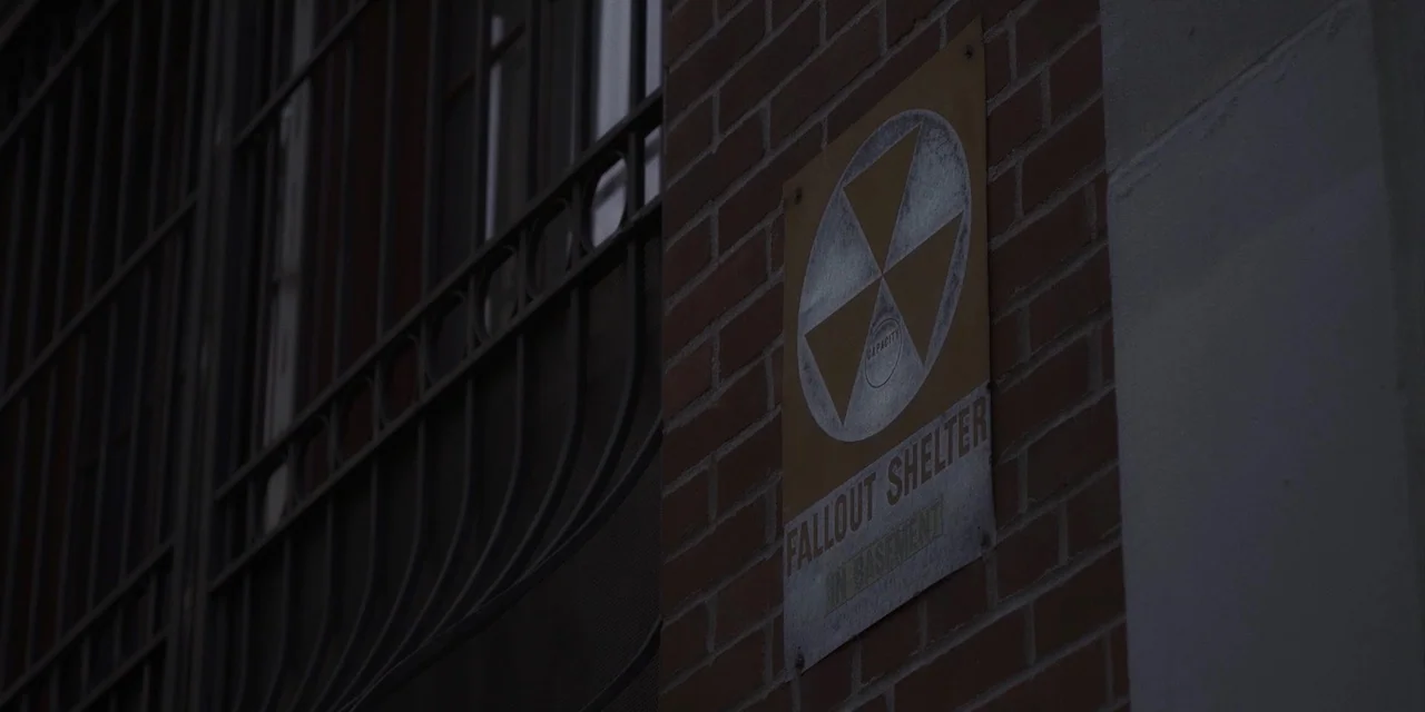 The Sound of Silence movie still: closeup — A red brick wall features a faded yellow metal fallout shelter sign mounted centrally. Black metal…
