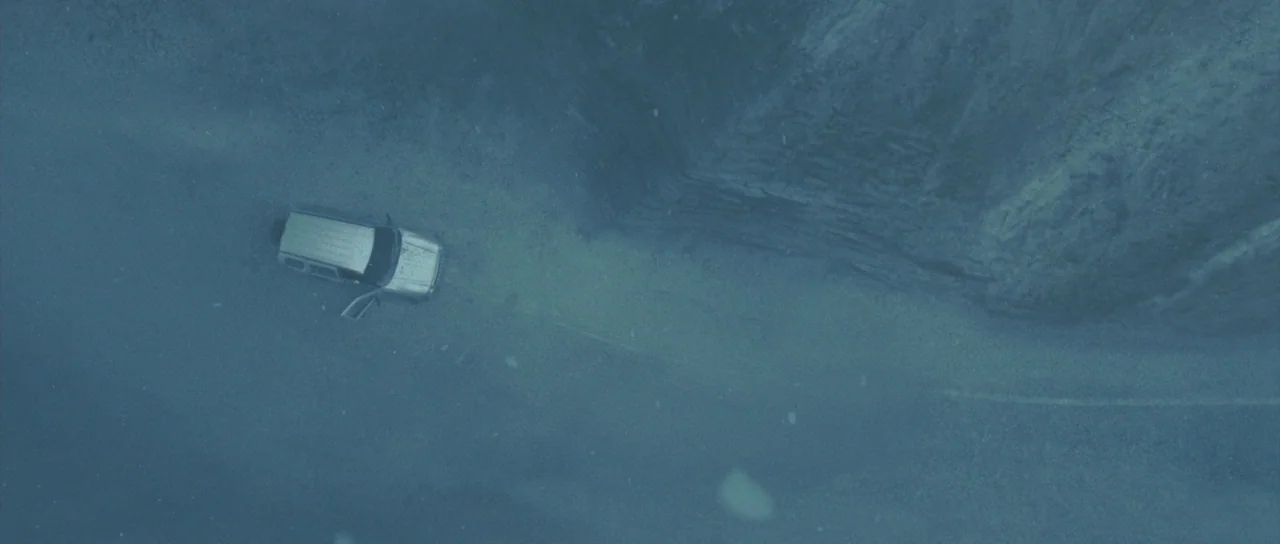 Silent Hill movie still: wide — A silver SUV is parked on a wet, gray asphalt road bordering a rock cliff. The scene is overcast and features light…