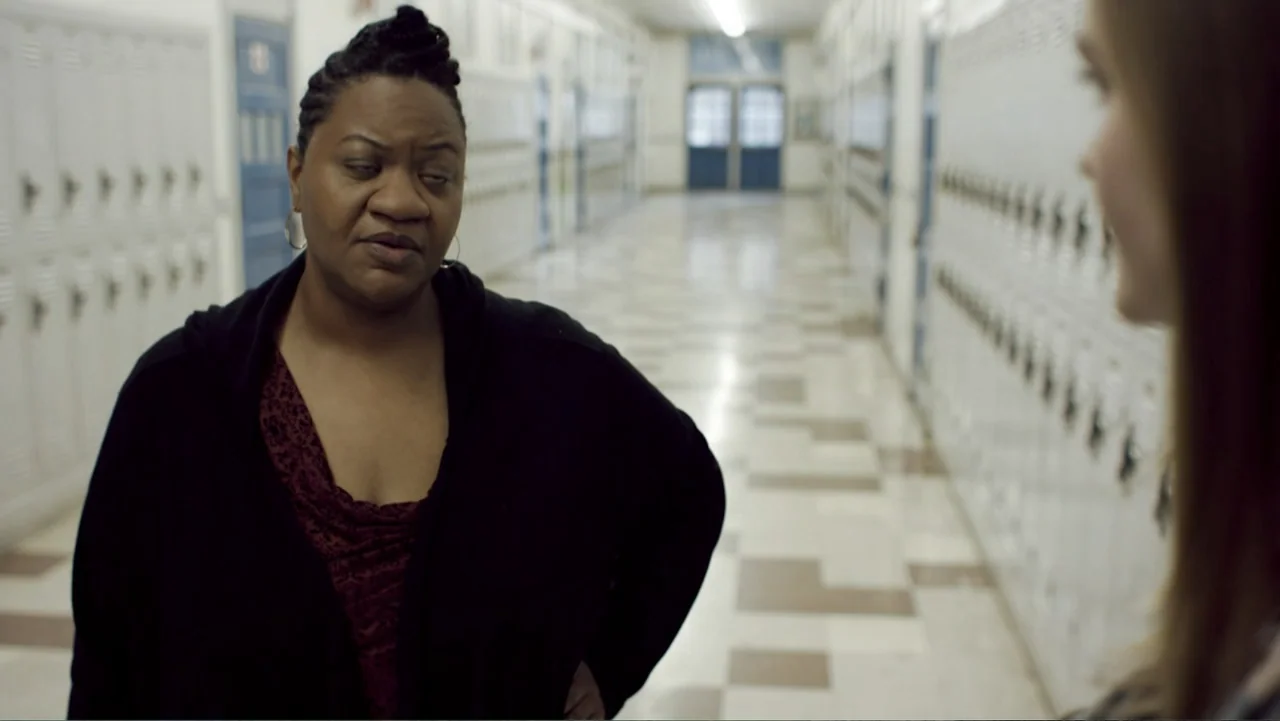 Totem (2022) movie still: medium — A long school hallway features rows of white metal lockers and checkered tile floors. A woman with braided hair…