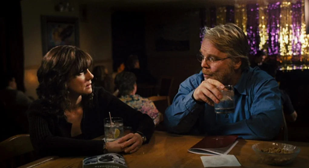 The Savages (2007) movie still: medium — A dimly lit bar features a man and woman seated at a wooden table. The man holds a glass while the woman…
